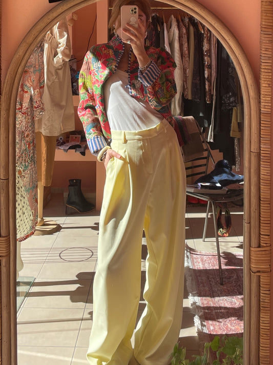 Person taking a mirror selfie wearing a colorful jacket and yellow pants in a room with clothes.